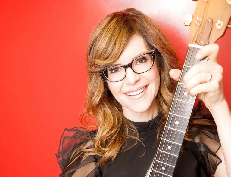 Lisa Loeb with guitar in front of a red backdrop. Juan Patino Photography
