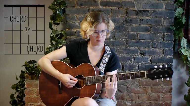 kate koenig, seated playing an acoustic guitar in front of a brick background with ivy